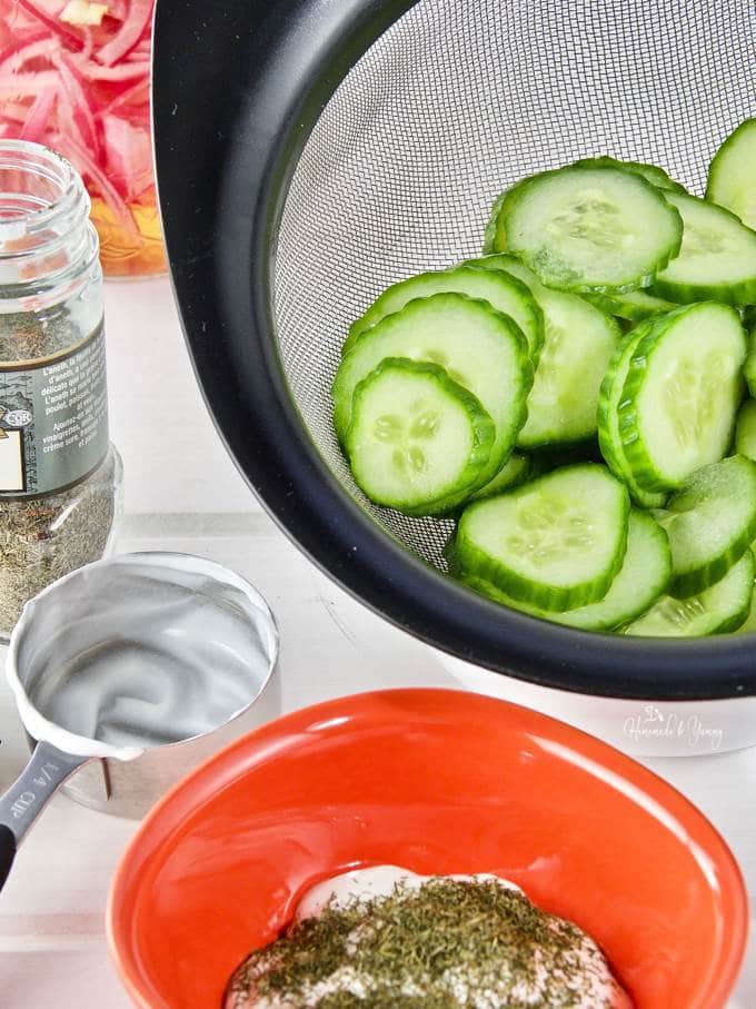 Cucumbers slices draining in a mesh strainer, salad ingredients off to the side.