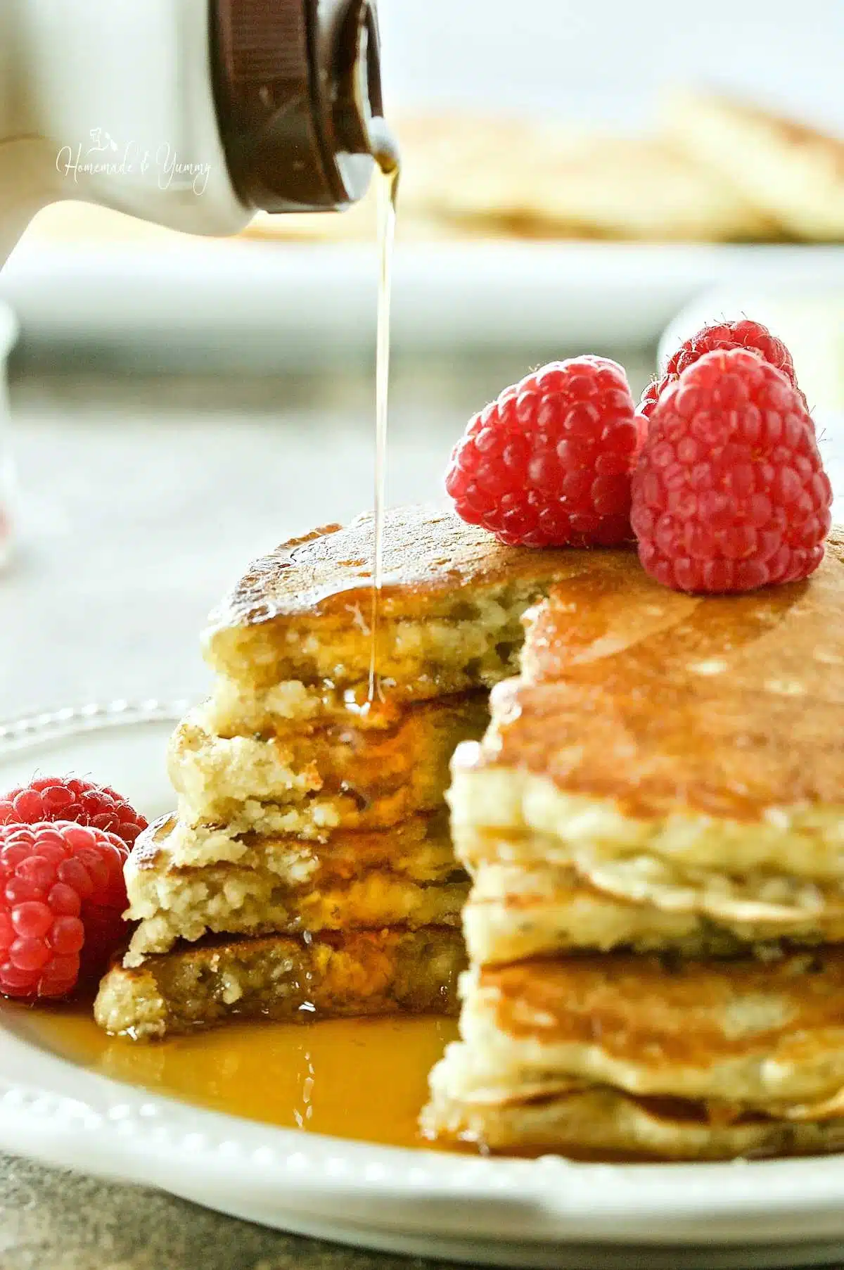 Syrup getting poured over a stack of oatmeal pancakes, topped with raspberries.