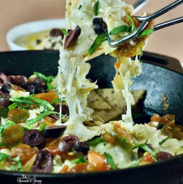 A pan of loaded Greek nachos, with a bowl of hummus in the background.